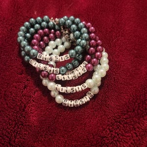 May include: A collection of four beaded bracelets featuring round, lustrous pearls in shades of dark teal, deep magenta, and white. Each bracelet incorporates white square letter beads spelling out either BIG SIS or LIL SIS in black text.