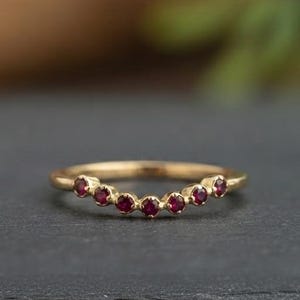 May include: A curved gold band ring featuring seven small, round, deep red gemstones set in a row along the top arc. The metal has a polished finish, and the design follows a gentle, symmetrical crescent shape across the finger band.