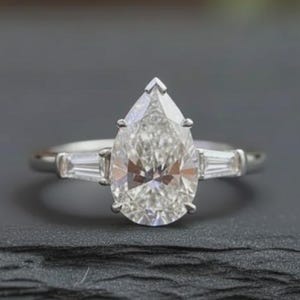May include: A silver ring with a large, pear-shaped diamond center stone and two smaller baguette diamonds on either side. The ring is set on a dark surface with a blurred background.