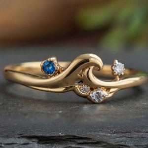 May include: A gold ring featuring a wave-shaped band design. The ring is accented with one small round blue gemstone and three clear white gemstones set along the curved, flowing metal structure of the band, creating a distinct ocean-inspired look.