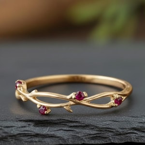 May include: A gold-colored ring with a delicate vine design. The ring features small, round, red gemstones set at intervals along the vine. The ring is displayed on a dark, textured surface.