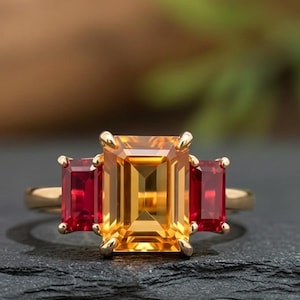 May include: A gold ring featuring a large, rectangular, yellow-orange gemstone flanked by two smaller, rectangular, red gemstones. The ring is set on a dark, textured surface, with a blurred background.