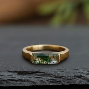 Może przedstawiać: Złoty pierścionek z prostokątnym, zielonym kamieniem mchu. Pierścionek leży na ciemnej, teksturowanej powierzchni. Kamień ma naturalny, zielony wzór. Tło jest rozmyte, z zielonymi i brązowymi tonami.
