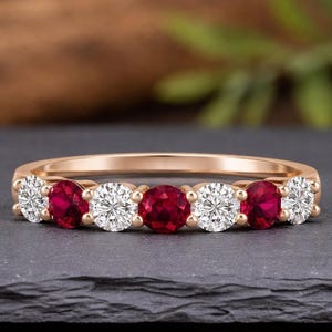 Könnte beinhalten: Ein roségoldener Ring mit abwechselnd runden Rubinen und Diamanten. Die Edelsteine sind in einer Reihe über der Oberseite des Bandes angeordnet. Der Ring wird auf einer dunkelgrauen Oberfläche präsentiert.