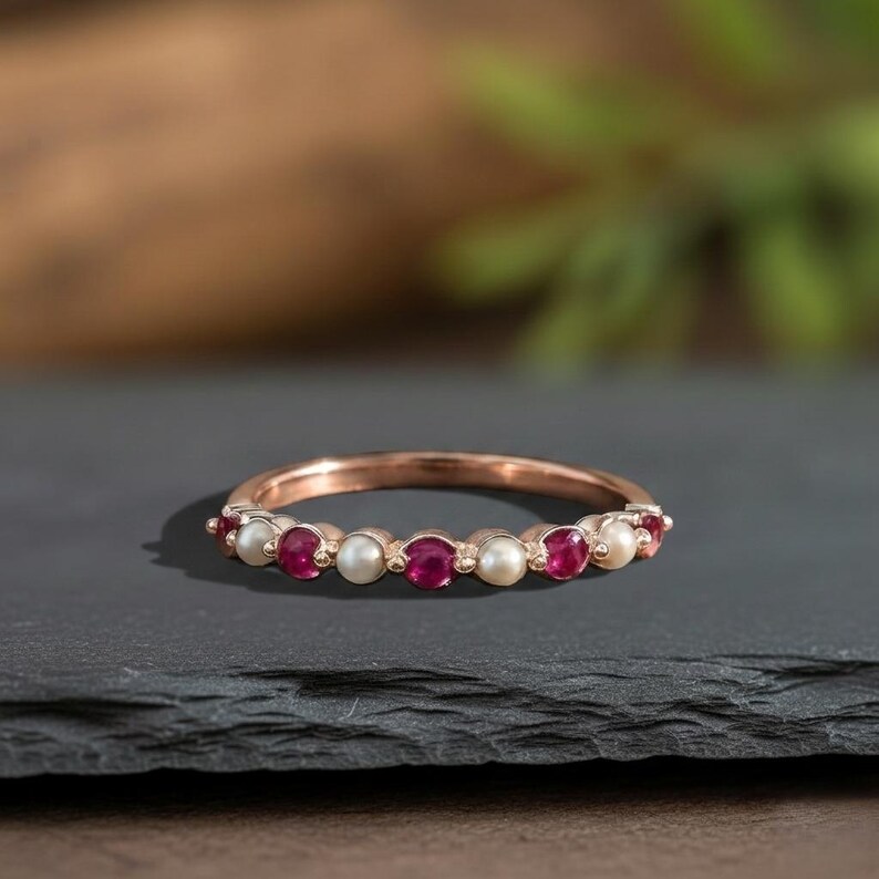 May include: A rose gold band ring featuring an alternating pattern of round, deep red rubies and small, creamy white pearls set in a continuous row along the top half of the metal band, creating a delicate and balanced jewellery piece.