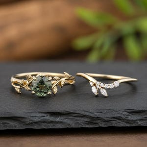 May include: A gold ring set featuring a round-cut olive green gemstone in a leaf-motif band alongside a curved gold band adorned with small clear stones arranged in a delicate branch pattern with marquise and round shapes.