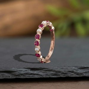 May include: A rose gold band ring featuring an alternating pattern of round, deep red rubies and small, white, spherical pearls set in a continuous row along the outer circumference of the metal band, creating a delicate and symmetrical jewellery piece.