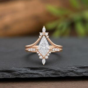 May include: Rose gold engagement ring with a marquise-cut diamond center stone and smaller accent diamonds. The ring features a unique crown-like design and a matching band, set against a dark background.