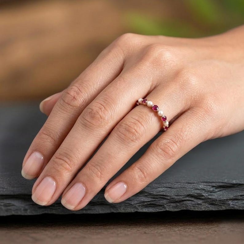 May include: A thin metal ring featuring an alternating pattern of small, round, white pearls and vibrant, circular, deep pink rubies set along the band. The jewellery piece is designed with a delicate, continuous arrangement of these gemstones.