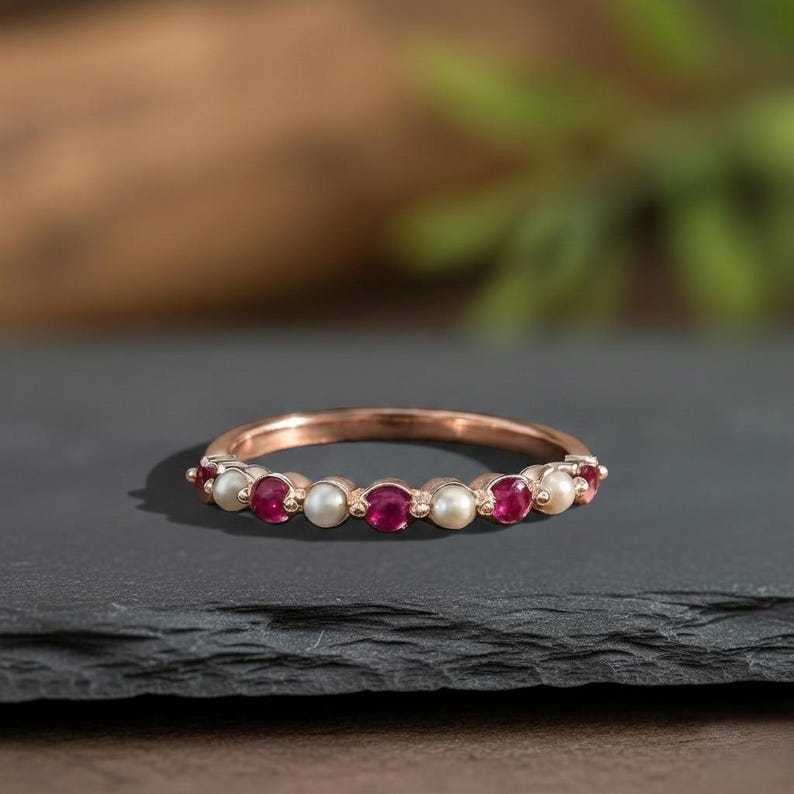May include: A rose gold band ring featuring an alternating pattern of round, deep red rubies and small, creamy white pearls set in a continuous row along the top half of the metal band, creating a delicate and balanced jewellery piece.