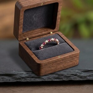 May include: A ring featuring alternating round red gemstones and white pearls set in a thin metal band, displayed inside an open dark brown wooden box with a grey velvet interior lining. The box has a hinged lid and a smooth, polished finish.