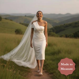 May include: A white, one-shoulder wedding dress with a knee-length hem. The dress features a draped accent on the shoulder and is paired with a long, flowing veil. The image is set outdoors with a grassy field and rolling hills in the background.