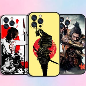 Può includere: Tre custodie per telefono con disegni a tema samurai. Una custodia presenta una grafica rossa e bianca di una donna con una spada. Un'altra mostra un samurai nero davanti a un sole rosso su sfondo giallo. La terza custodia mostra un'illustrazione dettagliata di un samurai.