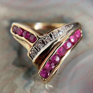 May include: A gold-toned ring with a V-shaped design. The ring features a row of small, round, deep red gemstones on each side of the V, and a row of small, clear gemstones in the center. The ring is set against a blurred background.