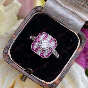 Peut inclure: Une bague de style vintage avec un diamant rond central, entouré d'accents de rubis rectangulaires dans un motif géométrique. La bague est sertie dans un anneau en argent ou en or blanc et présentée dans un écrin doublé de velours noir.