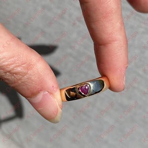 Peut inclure: Une bague en or avec une pierre en forme de cœur de couleur rubis. La bague est tenue entre les doigts, mettant en valeur la surface polie et le rouge vif de la pierre précieuse. L'anneau est lisse et brillant.