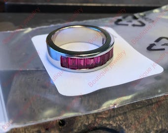 Könnte beinhalten: Ein silberner Bandring mit einer Reihe rechteckiger, leuchtend rosa Edelsteine. Der Ring liegt auf einer weißen Oberfläche. Im Hintergrund ist der Schriftzug "Boutique Jewellry" zu sehen.
