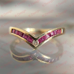 May include: A gold-toned ring with a V-shaped design. The ring is set with square-cut, vibrant red gemstones and a single, clear diamond. The ring's design is reflected in the shiny surface below.