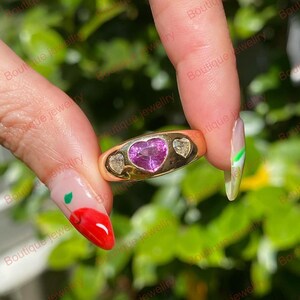 May include: A gold ring featuring a heart-shaped pink gemstone in the center, flanked by two smaller, clear gemstones. The ring has a smooth, polished band and is held up against a blurred green background.