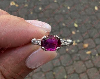Könnte beinhalten: Ein silberner Ring mit einem großen, oval geschliffenen, magentafarbenen Edelstein als Mittelpunkt. Der Ring hat kleinere, klare Edelsteine auf beiden Seiten des Hauptsteins. Das Band hat ein verziertes, strukturiertes Design.