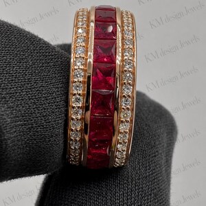May include: A rose gold band ring featuring a central row of square-cut deep red rubies flanked by two rows of small round brilliant-cut white diamonds. The ring is set against a dark gray fabric background with the text KM design Jewels visible.