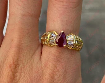 Könnte beinhalten: Ein goldener Ring mit einem birnenförmigen, tiefroten Edelstein in der Mitte, flankiert von Baguette- und runden, klaren Steinen. Der Ring hat ein breites Band mit zusätzlichen kleinen, klaren Steinen. Der Ring befindet sich an einem Finger.