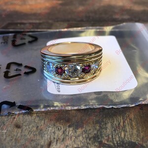 May include: A gold-toned ring with a band design. The ring features a row of clear and red gemstones. The ring is on a white surface.