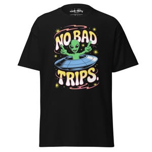 Könnte beinhalten: Schwarzes T-Shirt mit einem Cartoon-Alien in einem UFO, das den Text "NO BAD TRIPS" zeigt. Der Alien ist grün und zeigt das Peace-Zeichen. Das Design umfasst gelbe Sterne und rosa und gelbe Wirbel.