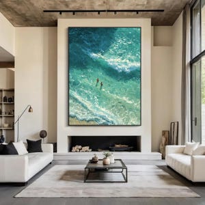 Puede incluir: Obra de arte enmarcada de gran tama&ntilde;o que representa una vista a&eacute;rea de las olas del oc&eacute;ano en tonos turquesa y blanco. Dos peque&ntilde;as figuras nadan en el agua cerca de la orilla. La obra se exhibe sobre una chimenea en un sal&oacute;n moderno.