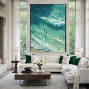 Puede incluir: Obra de arte enmarcada de gran tama&ntilde;o que representa una vista a&eacute;rea de las olas turquesas del oc&eacute;ano con dos figuras nadando. La pintura cuelga sobre un sof&aacute; seccional blanco con cojines verdes y blancos. Una mesa de centro de madera oscura est&aacute; frente al sof&aacute;.