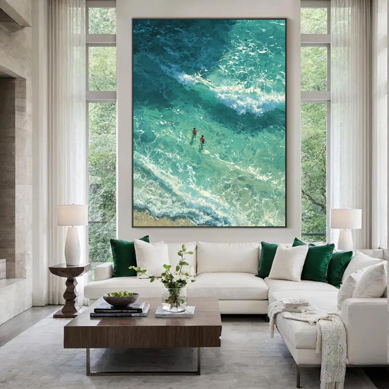 Puede incluir: Obra de arte enmarcada de gran tama&ntilde;o que representa una vista a&eacute;rea de las olas turquesas del oc&eacute;ano con dos figuras nadando. La pintura cuelga sobre un sof&aacute; seccional blanco con cojines verdes y blancos. Una mesa de centro de madera oscura est&aacute; frente al sof&aacute;.