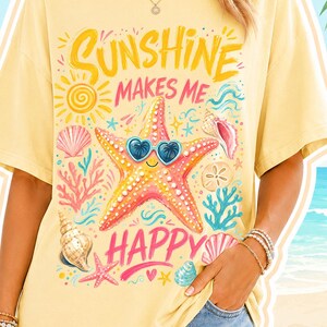 May include: A yellow t-shirt with the text "Sunshine Makes Me Happy" and a starfish wearing heart-shaped sunglasses. The shirt features a beach-themed design with seashells, coral, and a sun graphic. The person is wearing denim shorts.