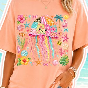 Puede incluir: Camiseta color melocotón con un diseño de medusa colorida con un patrón de patchwork, rodeada de ilustraciones con temática marina. El diseño incluye estrellas de mar, conchas marinas y flores tropicales. La camiseta se combina con pantalones cortos vaqueros.