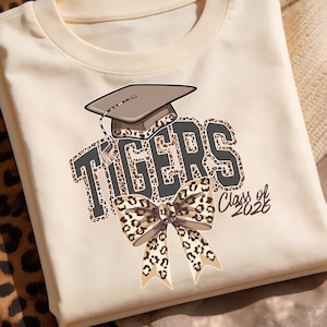 Puede incluir: Camiseta color crema con la palabra "TIGERS" en un diseño audaz de estampado de leopardo, coronada con un gorro de graduación. Un lazo con estampado de leopardo está debajo, con "Class of 2026" en blanco. La camiseta está doblada.
