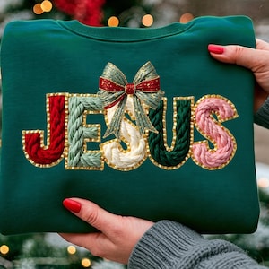 May include: A dark green sweatshirt with the word "JESUS" embroidered in a textured, colorful design. The letters are outlined in gold and feature red, green, white, and pink yarn. A decorative bow sits above the "E".