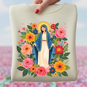 Op de afbeelding: Crèmekleurige sweatshirt met een afbeelding van een vrouw in een blauwe mantel en witte jurk, omringd door kleurrijke bloemen. Het ontwerp bevat een gouden halo, tegen een vage achtergrond.