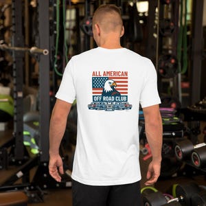 May include: White t-shirt with a graphic on the back. The graphic includes an American flag, an eagle, and the text "ALL AMERICAN OFF ROAD CLUB." Below the text are illustrations of off-road vehicles.