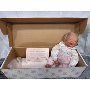 Könnte beinhalten: Eine schlafende Puppe mit blondem Haar und einem Schnuller im Mund, bekleidet mit einem weißen Langarmshirt mit rosa Blumenstickerei und einer rosa-weiß karierten Hose. Die Puppe liegt in einem Karton mit einem Echtheitszertifikat.