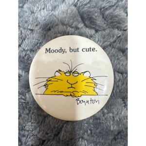 Op de afbeelding: Een ronde, witte knoop met de tekst "Moody, but cute." toont een cartoonkat met gele vacht en een humeurige uitdrukking. De illustratie van de kat is zwart omlijnd, met daaronder de handtekening van de kunstenaar.