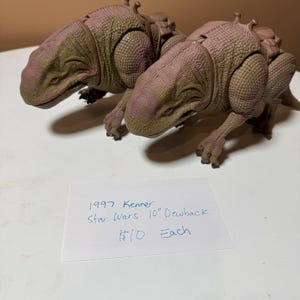 May include: Two 1997 Kenner Star Wars 10-inch Dewback figures. The figures are reptilian creatures with a textured, scaled appearance. The color scheme includes shades of brown, green, and pink. Each figure has a saddle-like structure on its back.