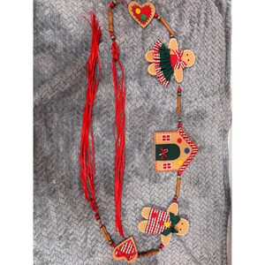 May include: A decorative garland featuring felt gingerbread-style figures, a house, and hearts strung together with cinnamon sticks and red ribbon. The ornaments are detailed with green and red plaid fabric, small buttons, and stitched accents.