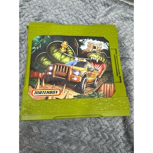 May include: A green Matchbox toy playset featuring a jungle scene with a yellow truck, a snake, and a dinosaur. The Matchbox logo is visible on the front of the playset.