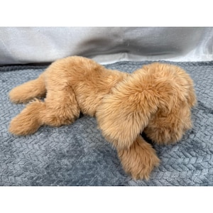 May include: A plush, golden-brown stuffed animal in a relaxed pose. The toy has soft, fluffy fur and is lying on a gray textured surface. The toy's limbs are extended, and its body is slightly curved.