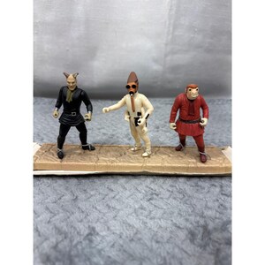 May include: Three Star Wars action figures. One figure is dressed in black with a horned head. The second figure is in white with a brown cone-shaped head and goggles. The third figure is in red with a monkey-like face. All figures are standing on a brown base.