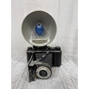 Può includere: Fotocamera vintage in bianco e nero con un accessorio flash. La fotocamera ha un design a soffietto pieghevole e un obiettivo con la scritta "FoldeX 20". Il flash ha un riflettore argentato e una lampadina blu.