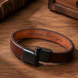 Puede incluir: Una pulsera de cuero marrón oscuro con un cierre de metal negro y el texto grabado Always love you en la superficie interior. La pulsera está fabricada con una banda de cuero liso con acabado mate y un mecanismo de cierre rectangular.