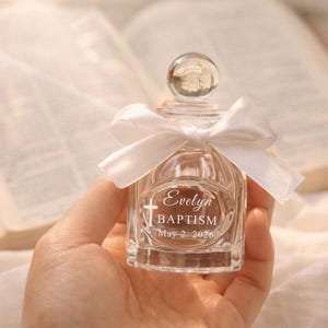 May include: A clear glass bottle with a spherical stopper and a white satin ribbon tied around the neck. The front of the bottle is etched with a cross symbol and the text: Evelyn, BAPTISM, May 2, 2026. The bottle has a square base.