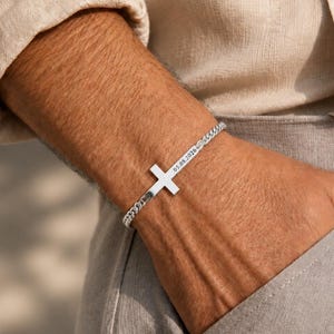 Puede incluir: Una pulsera de cadena de eslabones plateada con un dije de cruz central. La cruz tiene grabada la fecha 05.08.2026. La pulsera está construida con eslabones metálicos entrelazados y presenta un acabado pulido en toda su estructura.