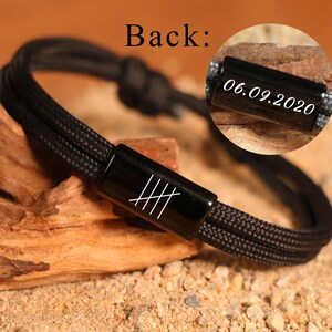 May include: Black braided bracelet with a rectangular black clasp featuring white hash marks. The back of the bracelet has the text "Back:" and a close-up shows the date "06.09.2020" engraved on a black clasp. The bracelet is on a wooden surface.