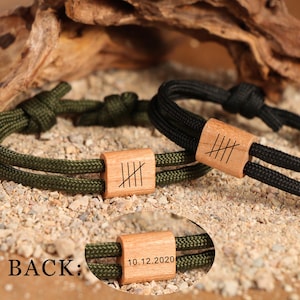 Könnte beinhalten: Nahaufnahme von zwei verstellbaren Seilarmbändern. Eines ist olivgrün, das andere schwarz. Jedes hat eine Holzperle mit eingeritzten Strichmarkierungen. Auf dem unteren Armband ist das Datum 10.12.2020 auf die Holzperle graviert. Das Wort "BACK:" ist ebenfalls sichtbar.